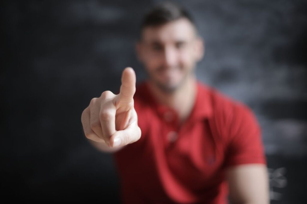 A man in a red shirt with a blurred face points his finger directly at the camera lens.